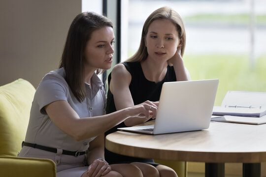 Two focused businesswomen, talking, discuss on-line project, learn collaborative task looking at laptop screen seated in modern office, mentor explaining new corporate software. Workflow, modern tech