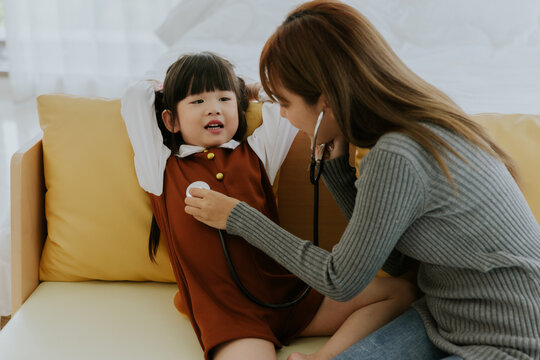 Young Asian Mother And Kid Girl Daughter With Stethoscope Sitting On Couch Role Play Doctor With Bear Doll Toy Together