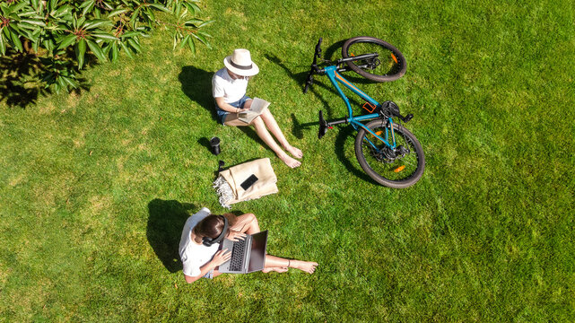 Young girls with bicycle in park, using laptop computer and headphones, two student girls studying online outdoors sitting on grass near bike, aerial drone view from above
