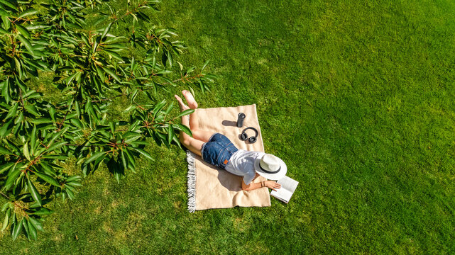 Young woman reading book in park, student girl relaxing outdoors sitting on grass with book and headphones, aerial drone view from above
