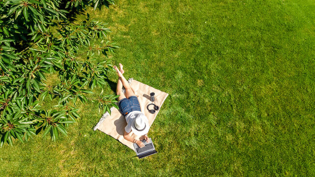 Young woman using laptop computer in park, student girl freelancer working and studying online outdoors sitting on grass with headphones and laptop, aerial drone view from above
