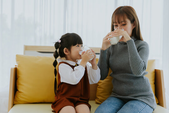 Young asian mother sitting on couch drinking milk with little kid girl daughter at home