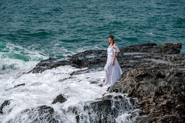 A woman stands on a rock in the sea during a storm. Dressed in a white long dress, the waves break on the rocks and white spray rises.
