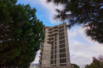 An apartment building under construction. Concrete structure with balconies and window openings. Pines ahead. Blue cloudy sky.