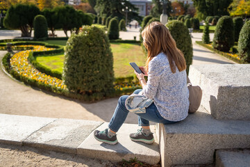 Woman sitting in a public park and looking at the mobile phone on a sunny autumn day.