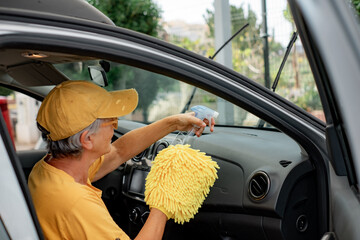 Smiling Caucasian senior woman in yellow t-shirt and hat cleans the car in a self-service car wash...