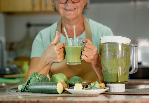 Mature Senior Woman Drinking Healthy Green Smoothie Holding A Glass With Straws. Smiling Caucasian Lady In Home Kitchen Drinking Detox Juice