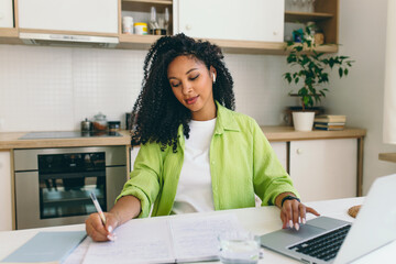 African american female in green shirt working at kitchen table, making to do-list, planning week. Brilliant student studying at home in front of laptop, noting down lecture listening in earphones