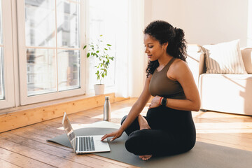 African american pregnant woman looking at laptop screen, searching for online workout tutorial or prenatal yoga sitting on floor on mat in sports wear, wearing smart watch to control heart rate - 543798111