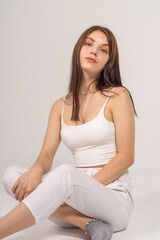 Attractive brown-haired girl in a white top and knitted trousers sitting posing on a white background