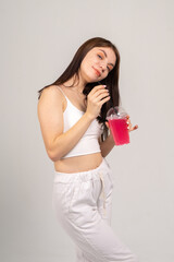 Attractive brown-haired girl in a white top and knitted trousers tries a pink cocktail through a straw on a white background