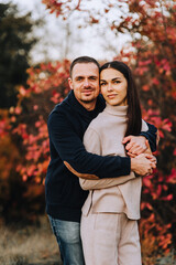 An adult smiling man and a beautiful young brunette woman are hugging, standing in nature in autumn, against the background of trees with red leaves. Photography, portrait.