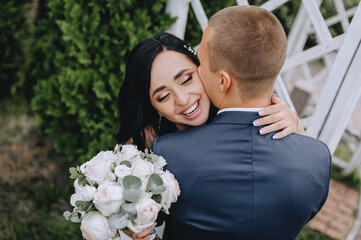 A beautiful, smiling brunette bride in a white dress with a bouquet of roses hugs the back of the groom in a blue suit. Wedding photography, close-up portrait.