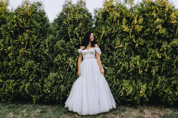 Fototapeta premium A beautiful bride in a white lush, lace dress with a bouquet in her hands stands near green thujas, cypress in the park, garden. Wedding photography, portrait.
