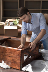 Serious man assembling wooden furniture on move-in day to own or rented flat, vertical view. Focused guy collects parts of nightstand table sit on floor in living room. Relocation, tenancy, new home