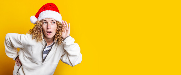 Curly-haired young woman in santa claus hat eavesdrops on a yellow background. Banner.