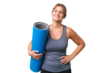 Middle-aged sport woman going to yoga classes while holding a mat over isolated background posing with arms at hip and smiling