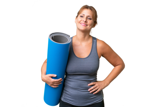 Middle-aged sport woman going to yoga classes while holding a mat over isolated background posing with arms at hip and smiling