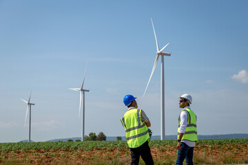 Two engineers wearing uniform and safety helmet work in wind turbine farm. Asian people engineers working at renewable energy farm, outdoor