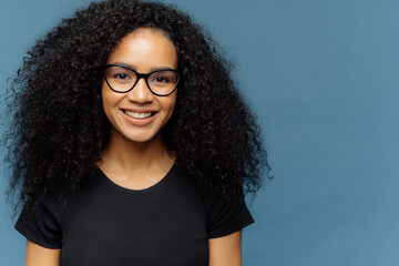 Headshot of pleasant looking dark skinned woman with Afro haircut, wears transparent glasses and...