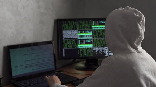Hooded IT programmer working on desktop computer in data center technical system control room. Young hacker working with computer, close up.
