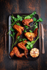 Place the roasted bbq chicken wings on a black baking sheet on a wooden background with parsley and garlic. Top view, flat layout