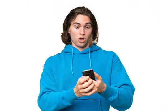 Young handsome man isolated on green chroma background looking at the camera while using the mobile with surprised expression