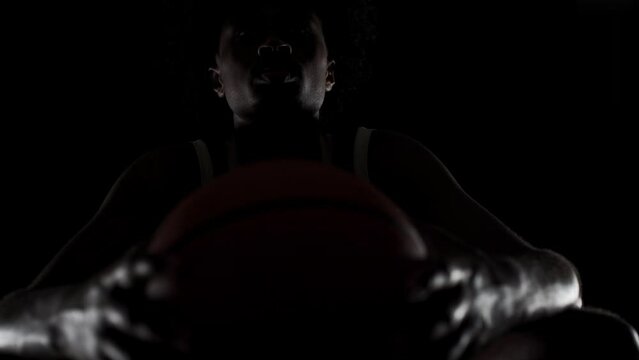 Sidelit basketball player silhouette sitting on a bench holding a ball. Concentrated african american sports man sidelit against black background.
