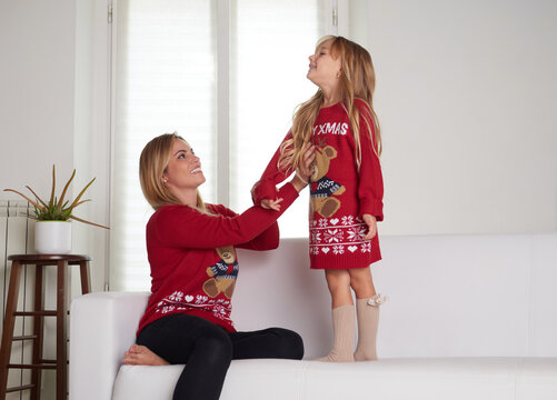 Young beautiful mother playing with daughter in livingroom dressing Christmas sweater