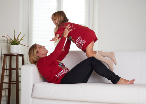 Young beautiful mother playing with daughter in livingroom dressing Christmas sweater