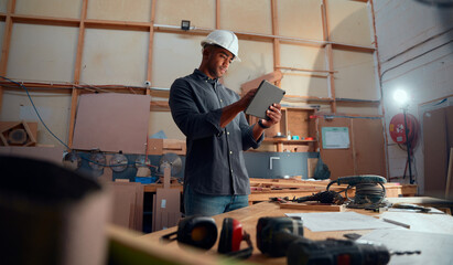 Young man in safety helmet using digital tablet by table with tools in woodworking factory