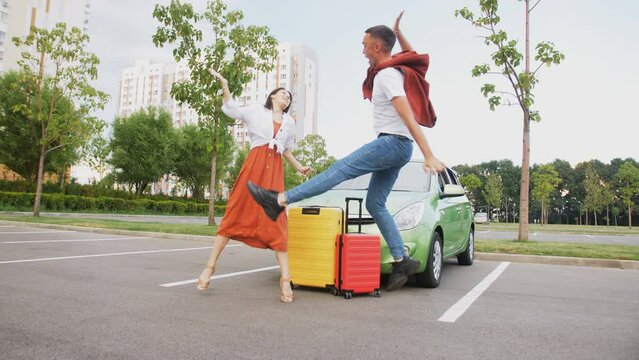 A guy in a jump slaps his girlfriend's palms with joy against the background of cars and colored suitcases. Lovers' journey.