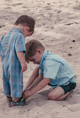 two adorable boy children, brother helping his younger brother with his shoes on sandy beach in...