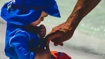 Grandfather's mature hand pointing at belly button of grandson, young toddler at the beach in...