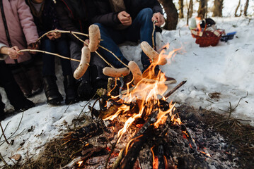 Family spending time together around campfire outdoors in the winter. People roasting sausages over the campfire using wooden sticks. Barbecue in nature.