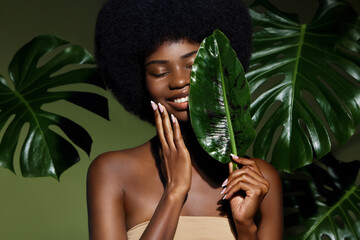 Organic cosmetic concept. Green beauty portrait. Young African American woman smiling and holding banana leaf against tropical green leaf. Natural cosmetics concept.