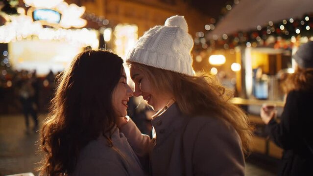 Two cheerful multi-racial girls hug and showing love, rubbing their noses in the middle of a busy New Year's Fair under the chimes of the New Year Christmas. Concept LGBT happiness freedom love