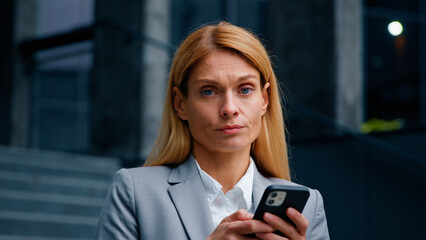 Fototapeta na wymiar Serious focused businesswoman holding phone standing in city sad frustrated woman receiving email message reading bad news loses in online bet upset about losing money feels unhappy negative emotions