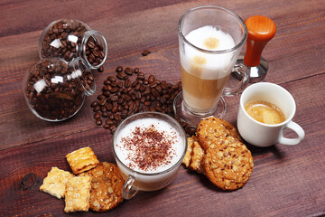 Breakfast with espresso, latte and cappuccino, crackers, cookies, holder with ground coffee, tamper and cans of coffee beans on wooden table.