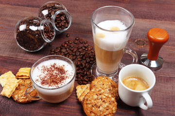 Breakfast with espresso, latte and cappuccino, crackers, cookies, holder with ground coffee, tamper and coffee beans on wooden table.