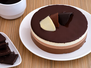 Chocolate mousse cake on a white plate. Plate with pieces of chocolate and cup of coffee near it on a bamboo napkin
