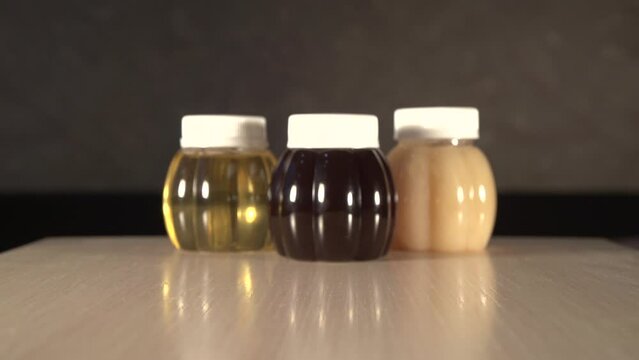 Close up zoom out of three plastic jars of different kinds honey on table with out of focus background. Light honey is Linden, dark honey is taiga, transparent honey is acacia. Jars with white caps