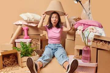 Shocked impressed Asian woman poses on floor under lampshade wears t shirt jeans and sneakers gathers personal belongings in cardboard boxes isolated over beige background. Relocation concept