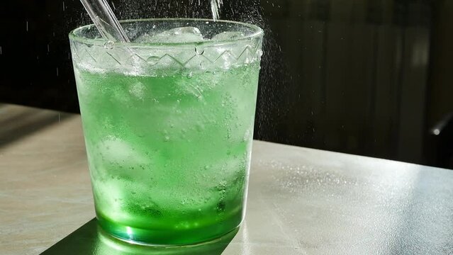Pouring green fizzy drink into a glass tumbler with ice cubes and a reusable glass straw in slow motion dolly shot. Flow and splash of tarragon or absinthe