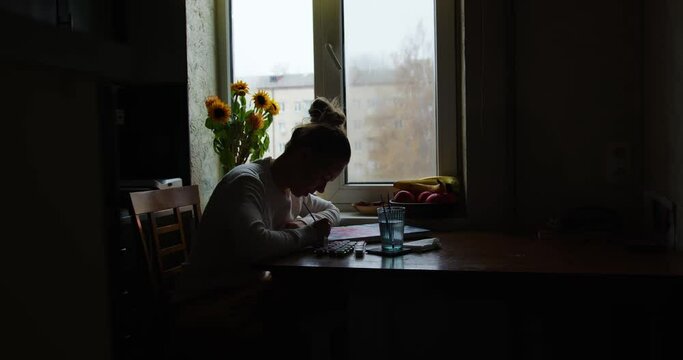 the girl draws a picture by numbers in the kitchen at the table opposite the window in the backlight. silhouette of a girl that paints a watercolor on canvas. overall plan