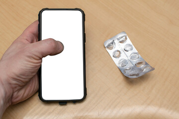 view of a smartphone with an isolated white screen in a person's hand against the background of a table on which a package of medicines and tablets, pills lies next to it