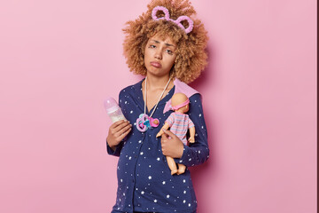 Expectant woman thinks about childbirth gets ready to become mother has pregnancy depression holds feeding bottle and doll dressed in pajama looks sadly at camera isolated over pink background