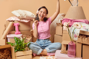 Joyful Asian woman dances and listens music via headphones sits crossed legs on floor prepares for relocation surrounded by personal belongings in carton boxes collects things. Moving Day concept