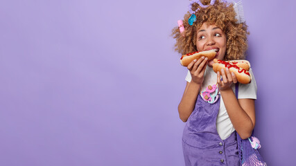 Fast food during pregnancy. Positive woman with curly bushy hair eats hot dogs with ketchup has...