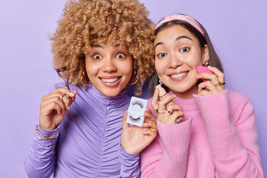 Two glad woman undego beauty procedures apply foundation hold eyelashes and curler smile gladfully make daily routines stand closely to each other against purple background. Domestic self care